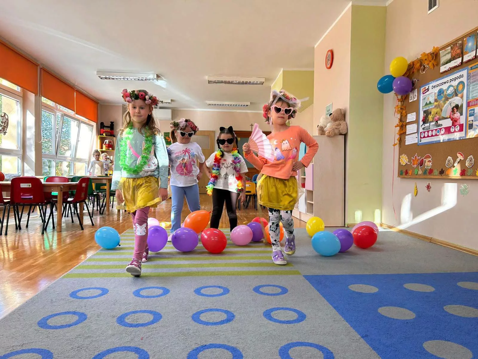 Cztery dziewczynki w strojach i okularach idą przez salę z balonami.